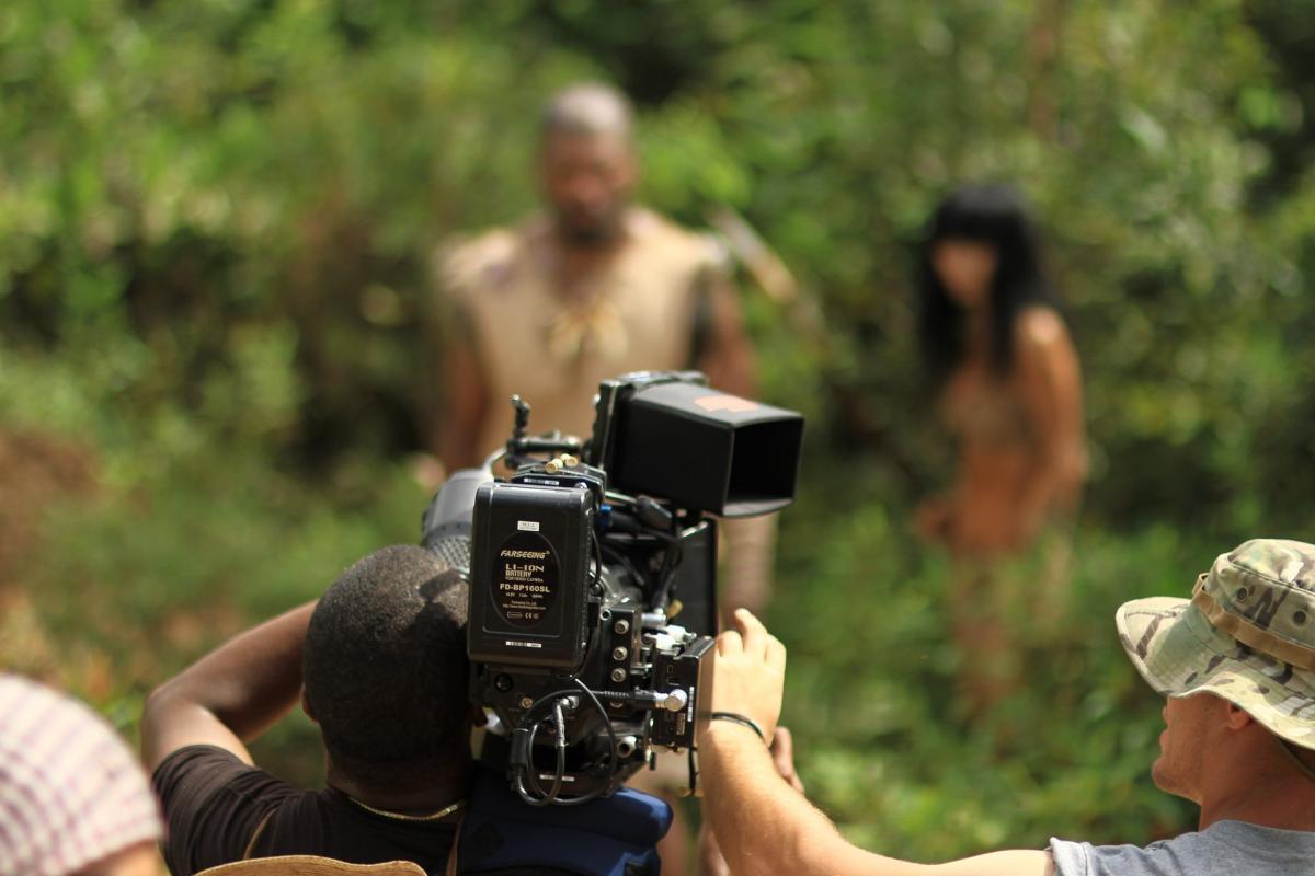 Un cámara rueda una serie, en la que aparecen actor y actriz al fondo.